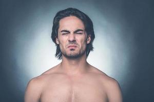 Feeling disgust. Portrait of young shirtless man keeping eyes closed and expressing negativity while standing against grey background photo