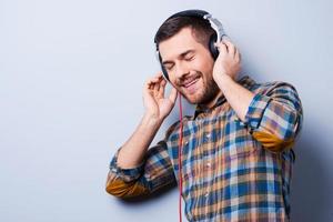 en las ondas de la música. un joven apuesto con auriculares que mantiene los ojos cerrados y sonríe mientras se enfrenta a un fondo gris foto