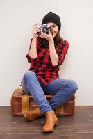 Give me a smile Beautiful young woman in headwear holding camera and looking through it while sitting on suitcase photo