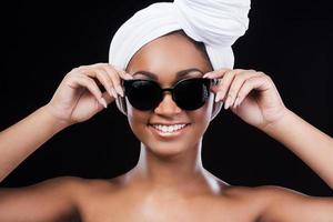 Beauty in style. Beautiful African woman wearing a headscarf and adjusting her sunglasses while looking at camera and standing against black background photo