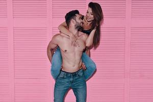 divirtiéndose. hombre guapo sin camisa que lleva a su novia sobre los hombros mientras está de pie contra el fondo rosa foto