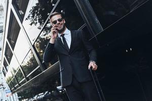 Good business talk. Handsome young man in full suit talking on the phone and smiling while standing outdoors photo