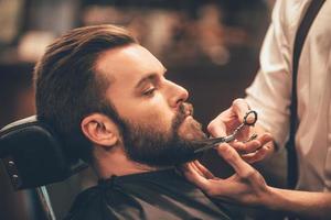 Getting perfect shape. Close-up side view of young bearded man getting beard haircut by hairdresser at barbershop photo