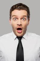 Outstanding news. Surprised young man in shirt and tie keeping mouth open and looking at camera while standing against grey background photo