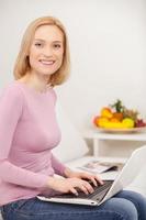 hacer negocios en casa. vista lateral alegre mujer de cabello rubio usando laptop y sonriendo a la cámara foto