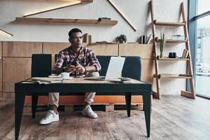 Searching for best answer. Thoughtful young man using his smart phone and looking away while sitting in the creative working space photo