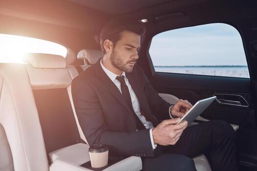 Considering the next step. Handsome young man in full suit working using digital tablet while sitting in the car photo