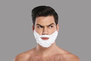 Ready to shave. Confused young man with foam all over his face looking away while standing against grey background photo