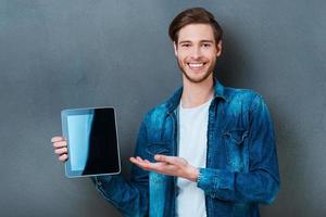 su anuncio en su tableta digital. un joven sonriente sosteniendo una tableta digital y apuntándola mientras se enfrenta a un fondo gris foto