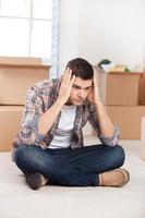 So many boxes to unpack. Depressed young man sitting on the floor and holding head in hands while cardboard boxes laying on the background photo