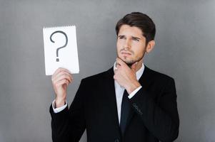In search of answer. Thoughtful young businessman holding note pad with question mark on it and looking away while standing against grey background photo