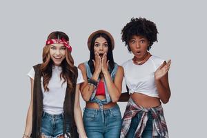 Three shocked young women looking at camera and making a face while standing against grey background photo