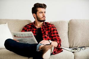 relajándose en el sofá. un joven pensativo sosteniendo un periódico y mirando hacia otro lado mientras se sienta en el sofá foto