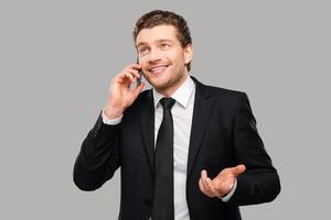 Good business talk. Portrait of confident young man in formalwear talking on the mobile phone and smiling while standing against grey background photo