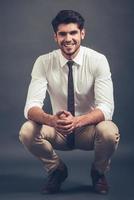 Elegant and cheerful. Full length of confident young handsome man looking at camera with smile while sitting crouched against grey background photo