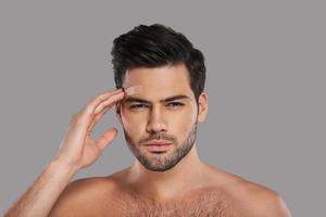 Feeling pain. Unhappy young man looking at camera and touching his adhesive bandage while standing against grey background photo