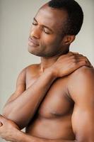 Feeling pain in shoulder. Young muscular African man touching his shoulder while standing against grey background photo