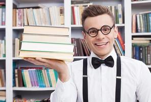 mis libros preferidos. un joven alegre con camisa y corbata de moño sentado en la mesa de la biblioteca y sosteniendo una pila de libros en la mano foto