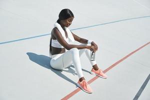 bella joven africana vestida con ropa deportiva sosteniendo una botella con agua mientras se sienta en una pista deportiva al aire libre foto