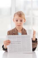 Shocking news. Surprised little girl in formalwear reading newspaper and holding a cup while sitting at the table photo