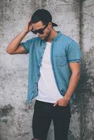 Casual style. Handsome young man in cap and sunglasses standing against the concrete wall photo