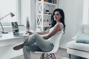 Surfing the net at home. Beautiful young woman using laptop and looking at camera with smile while sitting at the desk at home photo