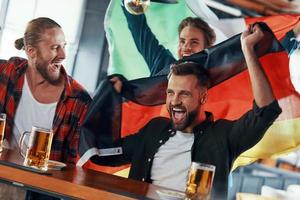 jóvenes cubiertos de banderas internacionales disfrutando de la cerveza mientras ven un partido deportivo en el pub foto