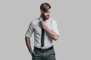 Perfect businessman. Young man in formalwear adjusting his necktie and looking away while standing against grey background photo