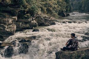 me quedaría aquí para siempre. apuesto joven moderno mirando hacia otro lado mientras se sienta en la roca cerca del río foto