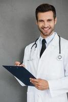 Checking the result. Happy young doctor in white uniform looking at camera and holding clipboard while standing against grey background photo