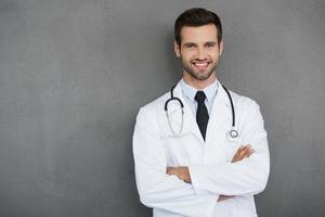 You can absolutely trust me. Confident young doctor in white uniform looking at camera and keeping arms crossed while standing against grey background photo