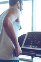 Exercising with great music. Rear view of young man in headphones running on treadmill in front of window at gym and holding his smart phone in his hand photo