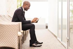 Checking his schedule on the go. Side view of cheerful young African businessman working on digital tablet while sitting on the chair photo