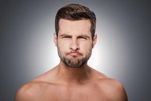 hombre haciendo muecas. retrato de un joven sin camisa mirando a la cámara y haciendo una mueca mientras se enfrenta a un fondo gris foto