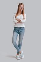 Confident woman. Full length of beautiful young woman in casual wear keeping arms crossed and looking at camera with smile while standing against grey background photo
