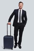 Time to travel. Full length of good looking young man with suitcase smiling and looking at camera while standing against grey background photo