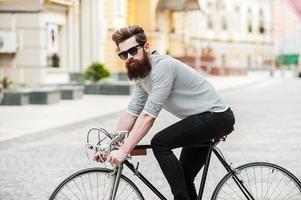 el camino sin tráfico vista lateral de un apuesto joven barbudo mirando a la cámara mientras monta en su bicicleta al aire libre foto