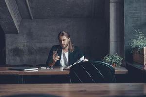 Staying connected.  Good looking young man in smart casual wear holding smart phone and looking at it while sitting at his desk in office photo