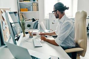 Creative businessman. Thoughtful young man in shirt working using computer and drinking coffee while sitting in the office photo