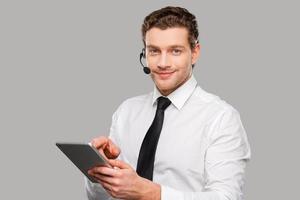 Always ready to help you. Handsome young man in formalwear and headset working on digital tablet while standing against grey background photo