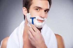 Handsome man shaving. Handsome shirtless young man shaving his face and looking at camera while standing isolated on grey background photo