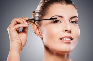 mujer haciendo maquillaje. hermosa mujer madura sosteniendo un cepillo de maquillaje en la ceja y sonriendo mientras se enfrenta a un fondo gris foto