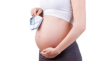 expectativa. imagen recortada de una mujer embarazada sosteniendo pequeños zapatos mientras está de pie aislada en blanco foto