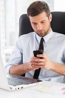Typing business message. Handsome young man in formalwear looking at his mobile phone while sitting at his working place photo