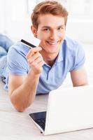 Home shopping. Handsome young man working on laptop and holding credit card while lying on the floor at his apartment photo