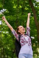 Happy traveler. Low angle view of happy young woman with backpack keeping arms raised and expressing positivity while standing in nature photo