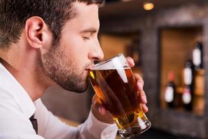 hombre bebiendo cerveza. vista lateral del apuesto joven bebiendo cerveza mientras está sentado en el mostrador del bar foto