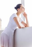 Thoughtful ballerina. Beautiful young ballerina holding hand on chin while looking through a window in ballet studio photo
