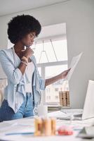 Beautiful young African woman analyzing fashion design sketch while standing near her desk in office photo