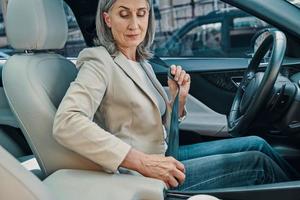 Mature beautiful woman in smart casual wear adjusting seat belt while sitting on front seat of the car photo